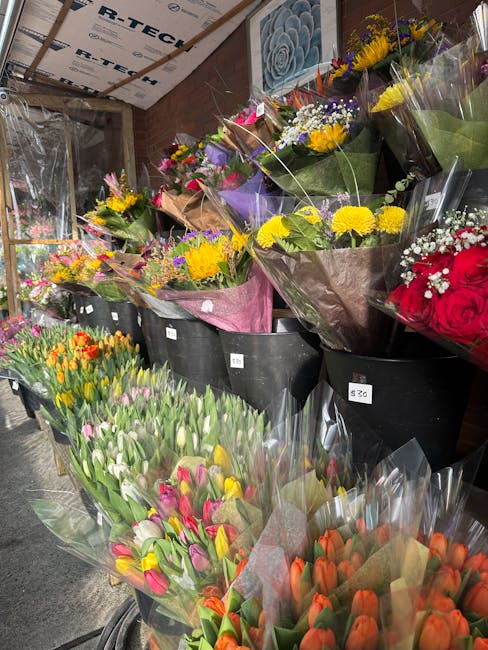 A vibrant display of fresh flowers arranged in large black buckets inside a flower shop. The bouquet arrangements feature a variety of colorful blooms, including yellow sunflowers, pink and white tuli