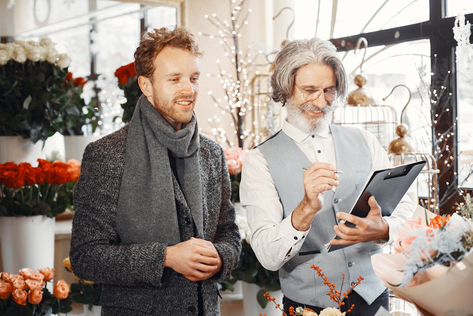 A young man with reddish-brown hair and a beard, dressed in a textured gray coat and matching scarf, stands inside a bright flower shop with natural light streaming through large windows. Beside him, 