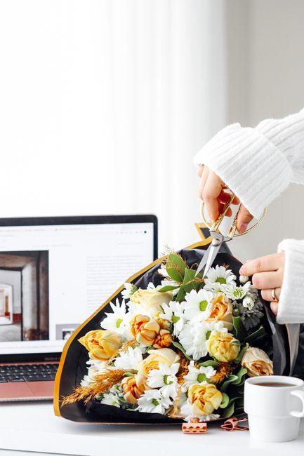 A professional florist's hand, wearing a white knitted sleeve, is seen using scissors to trim a bouquet of fresh flowers placed on a white surface. The bouquet features a vibrant mix of cream-colored 