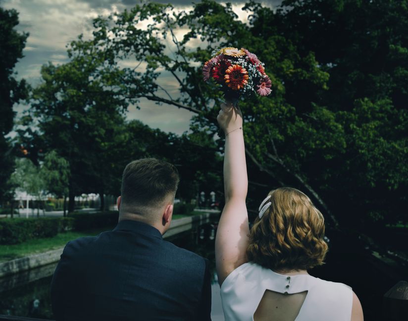 A woman with curly, shoulder-length reddish hair, wearing a white dress with a small back cutout, holds a vibrant bouquet high above her head during an outdoor event. The bouquet features a mix of lar