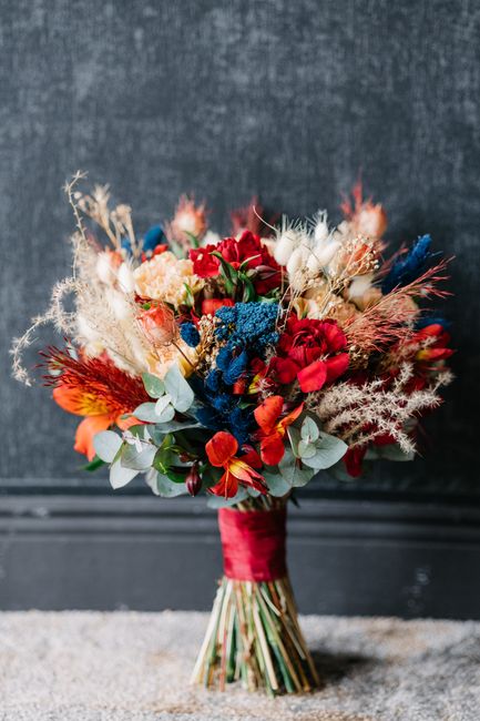 A vibrant bouquet featuring a mix of red, orange, white, and blue flowers arranged in a lush, textured display. The bouquet includes red roses, orange and white blooms, clusters of blue flowers, and s