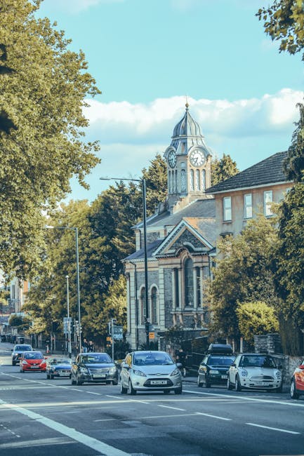 The image shows a street scene in New Barnet with a variety of parked and moving cars along the road, framed by lush green trees on both sides. In the background, there is a historic church with a pro
