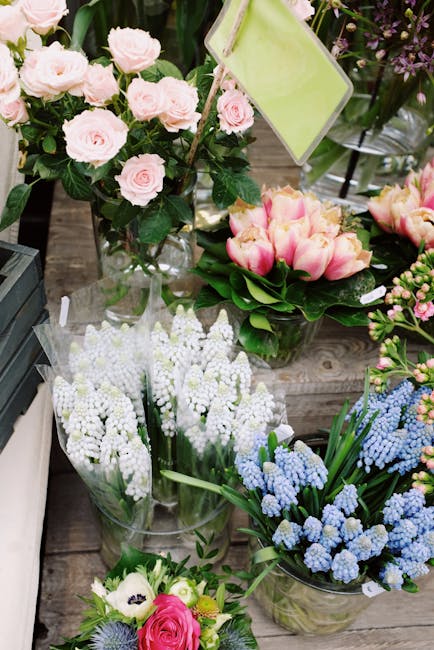 A floral display featuring an assortment of fresh flowers including pale pink roses arranged in a clear glass vase, surrounded by pink tulips in a round glass container with green foliage. Below, ther