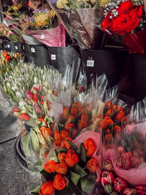 A display of various fresh flower bouquets arranged in black buckets at a florist shop, including a bouquet of vibrant orange tulips wrapped in pink tissue paper, a bouquet of pink and white tulips, a